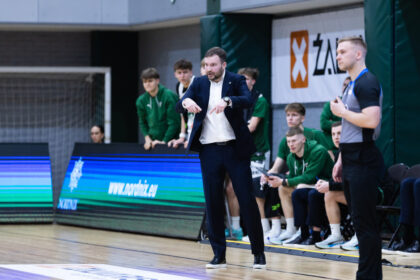Coach in a dark suit gives instructions to a team on the sideline of an indoor basketball court.