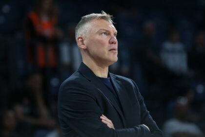 Man in a dark suit stands with arms crossed, looking upward in a dim arena.