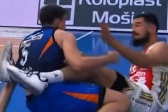Two male athletes grapple on a blue sports court; one in blue/orange with leg up, the other in white/red reaching toward him