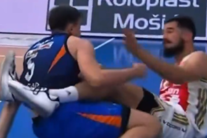 Two male athletes grapple on a blue sports court; one in blue/orange with leg up, the other in white/red reaching toward him