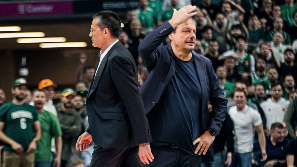 Two men in dark suits on a basketball court, one raising his hand as a cheering crowd watches behind them.