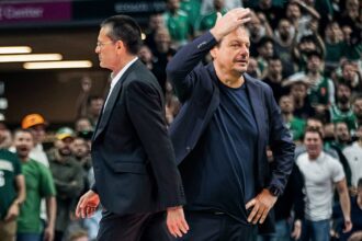 Two men in dark suits on a basketball court, one raising his hand as a cheering crowd watches behind them.