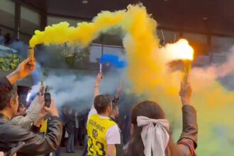 Crowd of fans outdoors waving colorful smoke flares (yellow and blue) during a celebration or protest at a stadium entrance.