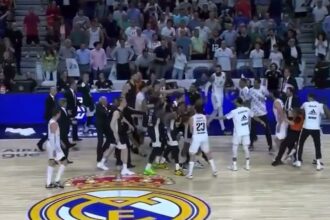 Basketball players gather in a celebratory huddle on a court with the Real Madrid emblem at center, surrounded by a packed audience in the stands.
