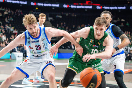 Basketball players in white/blue and green uniforms battle for the ball during a fast-paced game in an arena, crowd in background (number 23 visible).