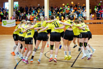 Women’s volleyball team in yellow jerseys form a circle, arms around each other, jumping in celebration on a court with a cheering crowd in the background.