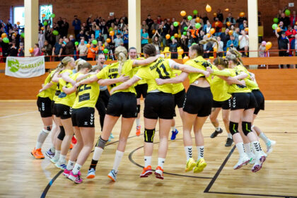 Women’s volleyball team in yellow jerseys form a circle, arms around each other, jumping in celebration on a court with a cheering crowd in the background.