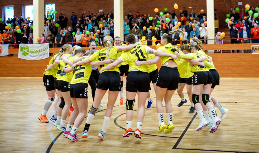 Women’s volleyball team in yellow jerseys form a circle, arms around each other, jumping in celebration on a court with a cheering crowd in the background.