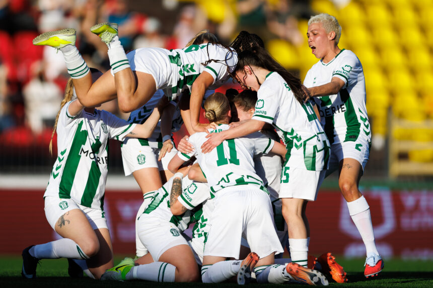 Female soccer players in white and green uniforms pile together in celebration on the field.