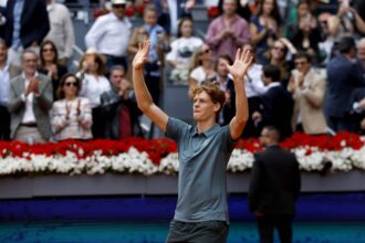 Man raises both arms in victory on a tennis court with a cheering crowd in the background amid red and white flowers.