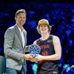 Presenter in a gray checked suit hands a basketball-shaped trophy to a smiling player in a black-and-red jersey and a white cap on a lit stage.