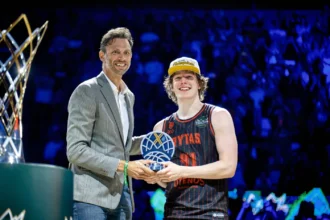 Presenter in a gray checked suit hands a basketball-shaped trophy to a smiling player in a black-and-red jersey and a white cap on a lit stage.