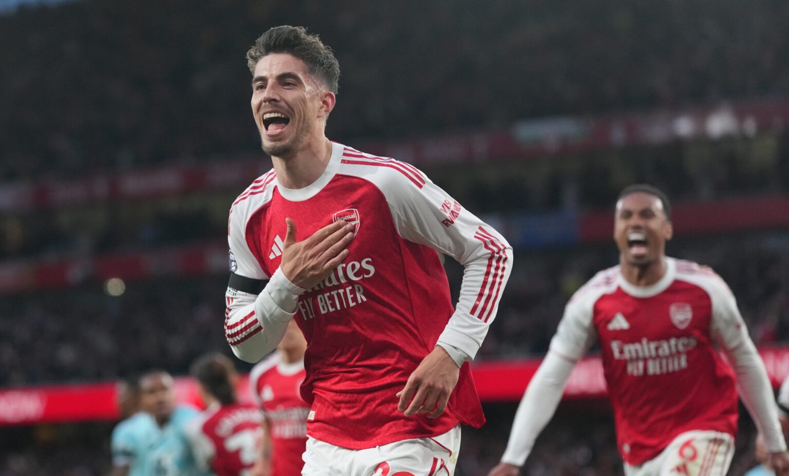 Arsenal players celebrate a goal on the pitch in red and white kits against a stadium backdrop.
