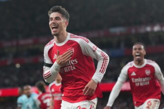 Arsenal players celebrate a goal on the pitch in red and white kits against a stadium backdrop.