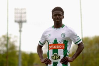 Soccer player in a white-and-green kit posing with hands on hips on a training field with stadium lights in the background, sponsor logos visible.