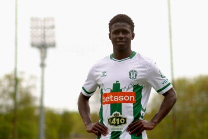 Soccer player in a white-and-green kit posing with hands on hips on a training field with stadium lights in the background, sponsor logos visible.