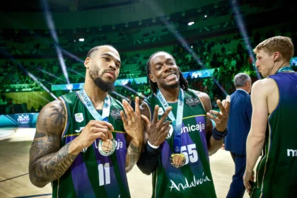 Three male basketball players in green uniforms pose with medals after a game on a lit indoor court, smiling as fans cheer in the green arena background.