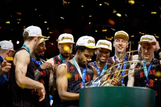 Basketball players in matching uniforms celebrate on stage with gold confetti and trophies.