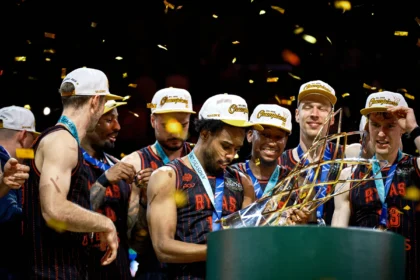 Basketball players in matching uniforms celebrate on stage with gold confetti and trophies.