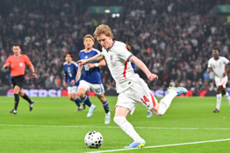 England player in a white jersey (#14) kicks the ball during a match as pursuing players and a crowd look on.