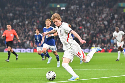 England player in a white jersey (#14) kicks the ball during a match as pursuing players and a crowd look on.