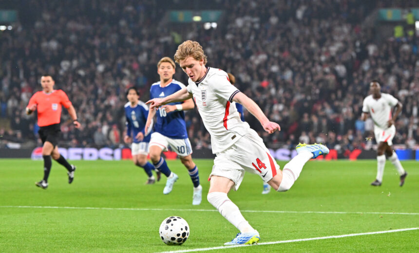 England player in a white jersey (#14) kicks the ball during a match as pursuing players and a crowd look on.