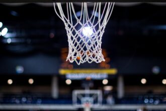 Close-up of a basketball hoop net hanging from the rim in a dark indoor arena, with a bright light behind it and a blurred backboard in the distance.