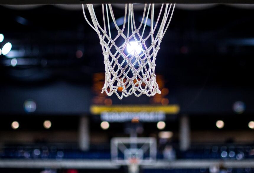 Close-up of a basketball hoop net hanging from the rim in a dark indoor arena, with a bright light behind it and a blurred backboard in the distance.