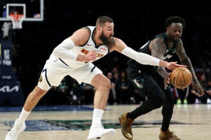 Two basketball players rush toward the ball on a glossy court, one in a white uniform reaching forward and the other in a dark uniform close behind.