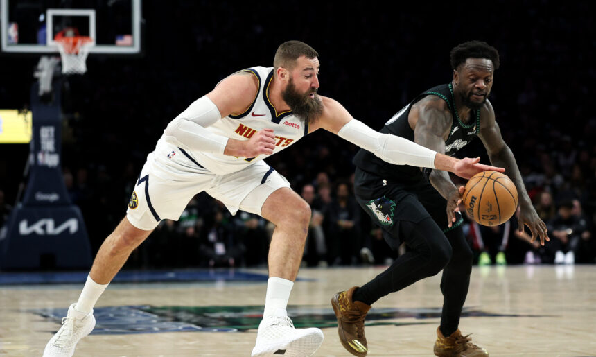 Two basketball players rush toward the ball on a glossy court, one in a white uniform reaching forward and the other in a dark uniform close behind.