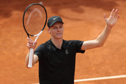 Tennis player on a clay court raises his right hand while holding a racket in his left hand, wearing a black shirt and cap.