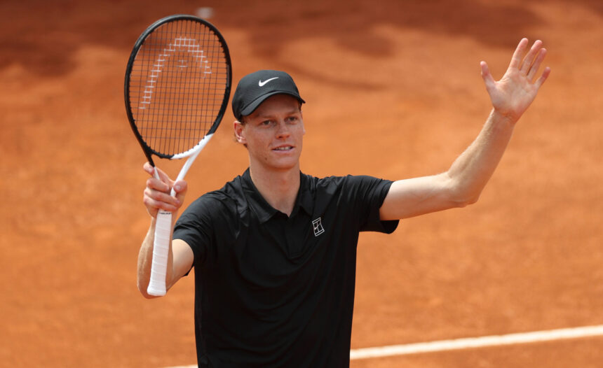 Tennis player on a clay court raises his right hand while holding a racket in his left hand, wearing a black shirt and cap.