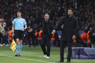 Football match sideline: referee in light blue kit with flag, and two coaches gesturing in front of a crowded stadium.