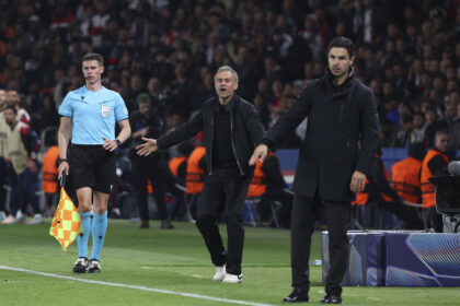 Football match sideline: referee in light blue kit with flag, and two coaches gesturing in front of a crowded stadium.