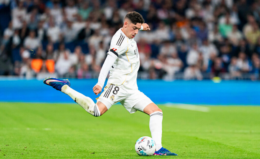 Soccer player in a white Real Madrid kit mid-kick on a green pitch, with a blurred stadium crowd in the background.
