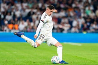 Soccer player in a white Real Madrid kit mid-kick on a green pitch, with a blurred stadium crowd in the background.