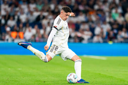 Soccer player in a white Real Madrid kit mid-kick on a green pitch, with a blurred stadium crowd in the background.