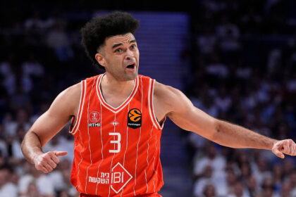 Basketball player in a red jersey with number 3, arms outstretched in mid-game action with a blurred crowd behind him.