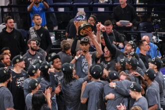 Basketball team in gray shirts celebrating, lifting a large trophy overhead.