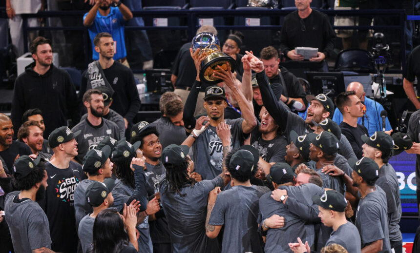 Basketball team in gray shirts celebrating, lifting a large trophy overhead.