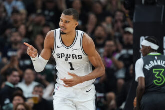 NBA player in a white San Antonio Spurs jersey dribbling on the court with a cheering crowd in the background, pointing with his right hand.