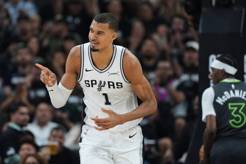 NBA player in a white San Antonio Spurs jersey dribbling on the court with a cheering crowd in the background, pointing with his right hand.