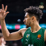 Basketball player wearing a green Žalgiris jersey raises two fingers in a victory gesture on the court.