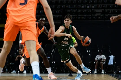 Basketball player in green jersey (number 35) dribbles past defenders on a court during a game, orange-clad opponents nearby.