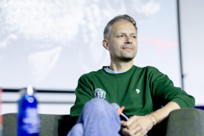 Portrait of a man with gray hair wearing a green sweatshirt, seated and looking to the right, with a blurred background and a Zalgiris badge on his chest.