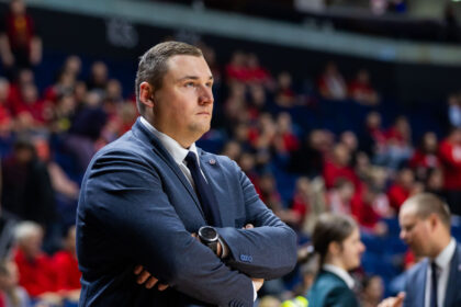 Man in a navy suit with arms crossed, standing in a crowded arena and looking focused on the event behind him.