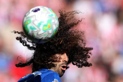 Soccer player in a blue jersey heads a multicolored ball mid-game with blurred stadium crowd in the background.