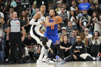 Knicks guard in blue jersey drives with the ball while a Spurs defender in white jersey guards him as fans watch from the stands in a packed arena.