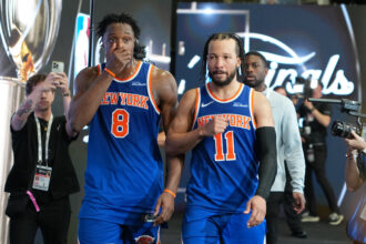 Two New York Knicks players in blue uniforms (numbers 8 and 11) walk through a media area with cameras and reporters nearby.