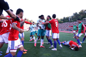 Soccer players in red and teal uniforms celebrate on a green field as fans fill the stadium stands behind them.